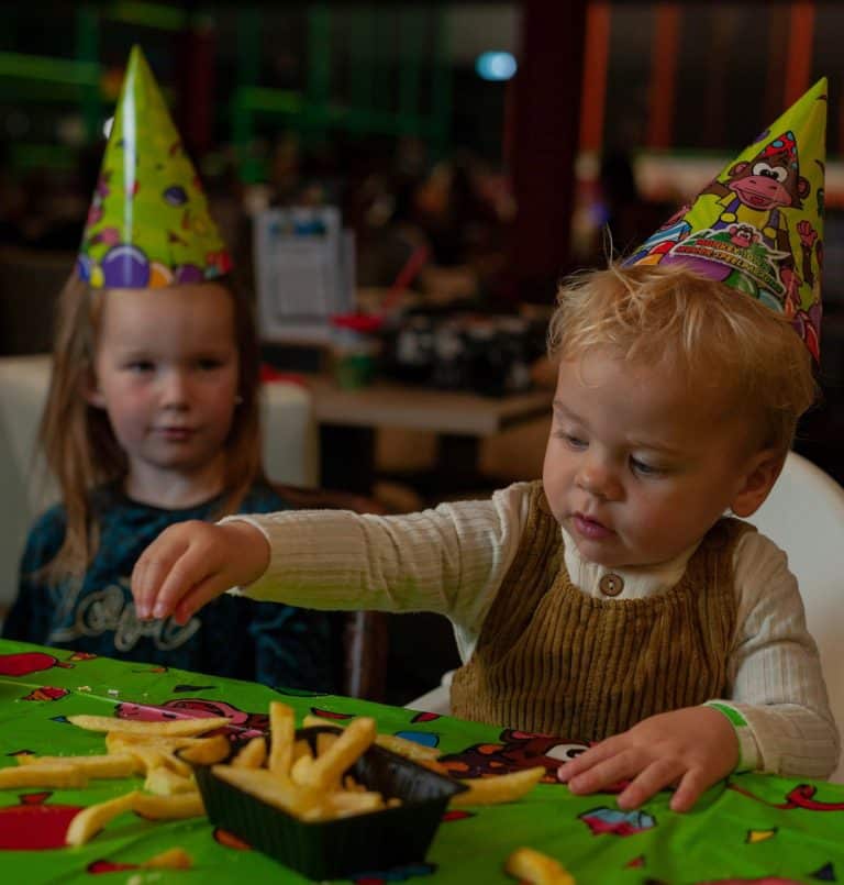 Jong kindje eet met zijn handjes een bakje patat. Het kind zit op een kinderstoel aan een tafel met een gekleurd tafelzeil en heeft een feestmuts op. Naast het kindje een meisje met lange haren en een feestmuts op die naar het kindje kijkt.