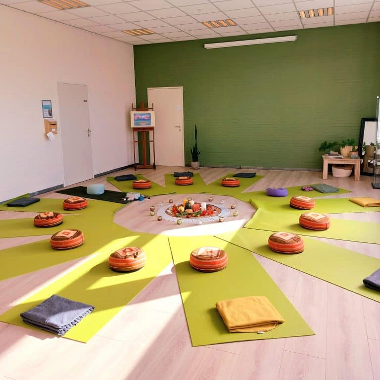 13 yoga matjes op de grond in een zaal. Op de matjes ligt een ronde kussen en een dekentje. In het midden staat een rustgevend tafereeltje met kaarsjes en kaarten.