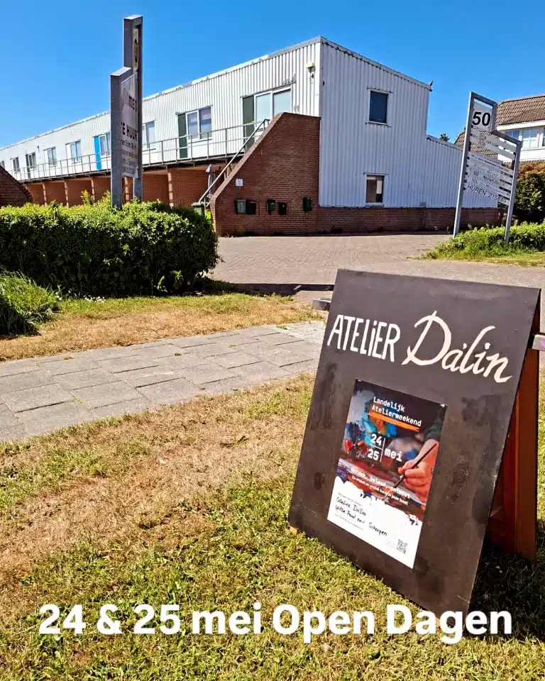 Buitenaanzicht van Atelier Da'Lin met op de voorgrond een bord waarop 'Landelijk aterlierweekend 24 en 25 mei ' staat, samen met afbeeldingen van een schilderpalet met een ahnd en verschillende kleuren verf en aanvullende tekst. Het moderne gebouw op de achtergrond heeft witte muren en grote ramen, en is omgeven door gras en struiken.