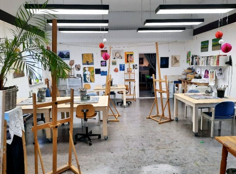 Interieur van een kunststudio met houten ezels, tafels en stoelen, omringd door schilderijen en tekeningen aan de muren. Er hangen kleurrijke papieren lantaarns aan het plafond en er staat een grote kamerplant links in beeld. Aan de rechterkant zijn planken met kunstmaterialen en boeken zichtbaar.