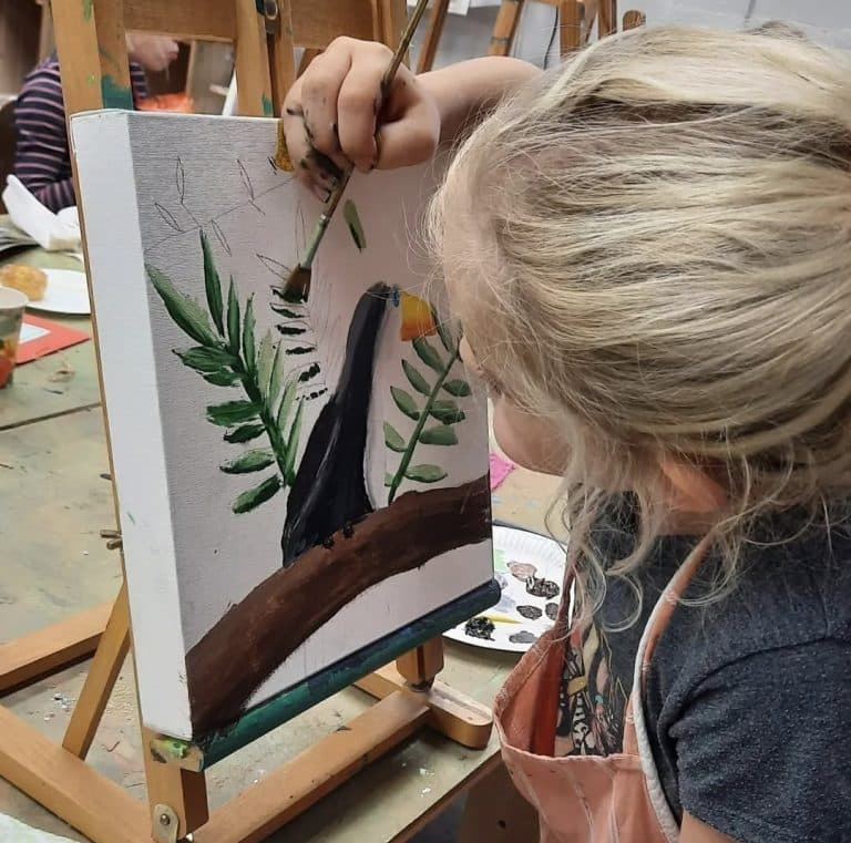 Kind schildert een zwart vogeltje met oranje snavel op een tak met groene bladeren, op een canvas op een schildersezel. Op de achtergrond zijn verfmaterialen, een tweede schildersezel en papieren bordjes met verf zichtbaar.