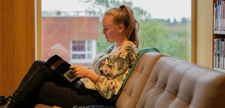 Jongedame leest een boek in bibliotheek. Ze zit op een bruine bank. Achter haar een stellingkast met boeken.